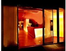 Ein modern gestalteter Hotelempfangsbereich mit überwiegend roten und warm beleuchteten Elementen. Ein beleuchteter Empfangstresen steht zentral, umgeben von gläsernen Türen und Vorhängen.