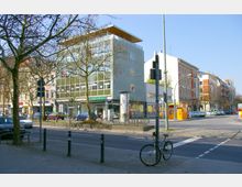 Straßenszene in einer Stadt mit moderner und historischer Bebauung; im Vordergrund ein Gehweg mit einem Fahrrad an einem Laternenmast, im Hintergrund ein vierstöckiges Gebäude mit Glasfassade sowie ältere Gebäude mit klassischen Fassaden. Straßenverkehr und parkende Autos sind sichtbar.