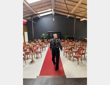 Person in einem Veranstaltungsraum mit Holzdecke und schwarzer Rückwand. Der Raum enthält rote Stühle mit Holzrahmen, kleine goldfarbene Tische und einen roten Teppich in der Mitte; im Hintergrund stehen Pflanzen und ein dunkler Schrank.