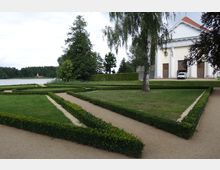 Ein geometrisch angelegter Garten mit niedrigen Buchsbaumhecken und bekiesten Wegen, im Hintergrund ein See mit bewaldetem Ufer. Rechts steht ein neoklassizistisches Gebäude mit weißen Säulen, großen Holztüren und einem roten Satteldach.