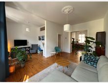 Helles Wohnzimmer mit Holzboden und weiß gestrichenen Wänden, möbliert mit einem grauen Sofa, einem runden Holztisch und verschiedenen Zimmerpflanzen. Im Hintergrund sind ein kleiner Arbeitsbereich mit Schreibtisch, ein Fernseher sowie ein Durchgang zu einem weiteren Raum mit Esstisch und großen Fenstern zu sehen.