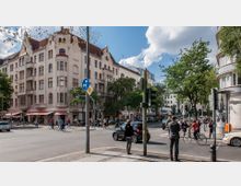 Straßenecke in einer Stadt mit einem mehrstöckigen Altbau mit heller Fassade und roten Dächern im Hintergrund, Geschäftszeilen mit Markisen im Erdgeschoss sowie Fußgängern, Radfahrern und Autos im Straßenverkehr. Im Vordergrund befinden sich Verkehrsampeln, Straßenschilder und Gehwege mit einzelnen Passanten.