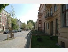 Straßenansicht in einem Wohngebiet mit historischen Mehrfamilienhäusern im Gründerzeitstil, die gelb und rot verputzt sind. Entlang der gepflasterten Straße stehen geparkte Autos, und auf der linken Straßenseite sind junge Bäume und ein schmaler Gehweg zu sehen.