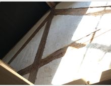 Fachwerk-Innenansicht mit weißen Wänden und sichtbaren Holzbalken, durch die Licht von außen fällt. Die Balkenstruktur ist diagonal und vertikal, mit Licht- und Schatteneffekten auf der Wand.