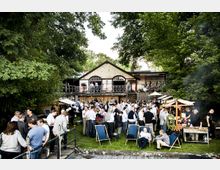 Ein Gartenfest vor einem historischen Gebäude mit Holzveranda und großen, gewölbten Fenstern; zahlreiche Personen stehen und sitzen auf dem Rasen, teils an Essensständen, während Sonnenschirme und Bäume für Schatten sorgen.