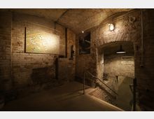 Ein historischer Keller mit freiliegenden Ziegelwänden und Gewölbedecken, beleuchtet von warmem Licht. An der Wand hängt eine Karte, und ein schmaler Gang unter einem Bogen führt tiefer in den Keller, begleitet von einem Schild mit der Aufschrift „Sperrgebiet“.