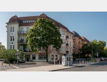 Ecke eines Wohngebäudes mit vier Stockwerken und hellem Putz, Ziegeldach und mehreren Balkonen; im Erdgeschoss befinden sich Geschäfte, darunter ein Beauty-Salon. Ein großer Baum steht vor dem Gebäude, daneben Gehwege, geparkte Autos und Straßenschilder.