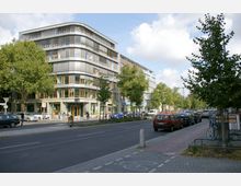 Moderne Stadtstraße mit einem fünfstöckigen Eckgebäude, das große Fenster und Balkone umfasst. Im Vordergrund Geh- und Radwege, geparkte Autos und vereinzelte Bäume entlang der Straße.