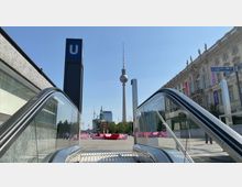Blick aus einer U-Bahn-Rolltreppe auf den Berliner Fernsehturm im Hintergrund, umgeben von modernen und historischen Gebäuden. Rechts im Bild ist ein klassizistisches Gebäude zu sehen, links die U-Bahn-Station mit einem großen blauen "U"-Schild.
