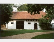 Ein einfaches Gebäude mit weiß verputzten Wänden, einem roten Ziegeldach und Holzdetails, darunter ein großes Doppeltor und eine kleinere Tür. Das Gebäude steht in einer grünen, baumreichen Umgebung mit einem geschwungenen Kiesweg im Vordergrund.