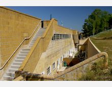 Flaches Gebäude mit gelblichen Klinkerwänden und einer breiten Treppe, die entlang der Außenwand verläuft. Im unteren Bereich befinden sich Fenster und eine Terrasse mit Sonnenschirmen, während im Hintergrund Bäume und ein klarer blauer Himmel sichtbar sind.