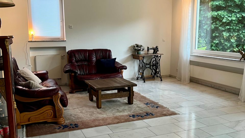 Helles Wohnzimmer mit großformatigem, hellgrauem Fliesenboden, zwei dunklen Ledersofas und einem niedrigen Holztisch auf einem Teppich. Große Fenster mit weißen Vorhängen und Heizkörpern darunter sowie ein kleiner Beistelltisch mit Nähmaschinenuntergestell stehen an den Wänden.