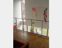 Innenraum eines modernen Gebäudes mit Holzboden und einer Galerie mit Metallgeländer. An den Wänden hängen großformatige, bunte Gemälde, und durch große Fenster sind ein Garten und Bäume sichtbar.