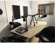 Ein Aufnahmestudio mit einem Holztisch, auf dem mehrere Mikrofone an schwenkbaren Armen befestigt sind, ein Audio-Mischpult und ein Paar Kopfhörer. Im Hintergrund sind schallabsorbierende Wandpaneele und ein Fenster zu einem angrenzenden Raum sichtbar.