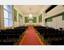 Ein klassischer Seminar- oder Vortragssaal mit grünen Wänden, weißen Holzpanelen, großen Fensterfronten mit grauen Vorhängen und einer zentralen roten Teppichbahn. Im Vordergrund stehen schwarze Stuhlreihen, und im Hintergrund ist eine erhöhte Bühne mit Holztischen und einer dekorativen weißen Türumrahmung zu sehen, die von einem Kronleuchter beleuchtet wird.