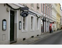 Straßenszene mit historischen Gebäuden, darunter ein Restaurant mit der Aufschrift „Sophien 11“ und dekorativen Girlanden. Die Fassaden sind in hellen Farben gehalten, teils mit Blumen geschmückt, und die Straße ist mit Kopfsteinpflaster befestigt.