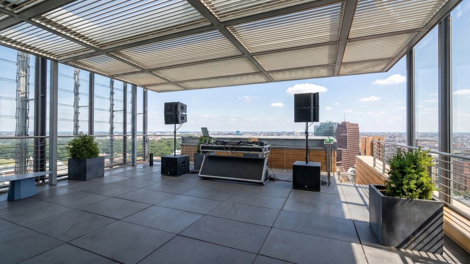 Rooftop terrace with a slatted metal canopy, glass and metal railings, and large floor tiles overlooking a city skyline. A DJ booth with speakers and cables is set up near the edge, with planter boxes containing shrubs at the corners.