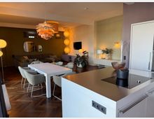 Open-plan modern kitchen and dining area with a white island countertop and built-in cooktop, leading to a dining table with upholstered chairs and a lounge with a sectional sofa and wall-mounted TV. The space features dark herringbone wood flooring, pendant and globe lighting, muted green and beige walls, and decorative mirrors and plants.