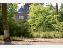 Backsteinhaus mit Dachgauben hinter einem Metallzaun, umgeben von dichtem grünen Baum- und Buschbestand in einem parkähnlichen Gelände. Im Vordergrund ist ein sandiger Weg mit einem Baum sichtbar.