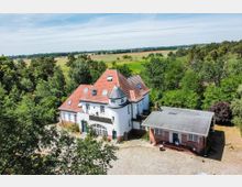 Großes weißes Haus mit roten Ziegeldächern und einem runden Turm, umgeben von einer gepflasterten Einfahrt, Bäumen und Wiesenlandschaft im Hintergrund. Neben dem Hauptgebäude steht ein flaches Nebengebäude mit Flachdach.