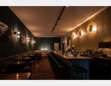 Eine gemütliche Bar mit dunklen Holztischen und -stühlen auf der linken Seite und einer langen, beleuchteten Theke mit Barhockern auf der rechten Seite. Die Wände sind dunkel gestrichen, mit moderner Wandbeleuchtung und einem warmen, gedämpften Licht, das eine intime Atmosphäre schafft.