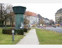 Ein Grünstreifen mit einem gepflasterten Weg und einem belüftungsartigen, grünen Turm in der Mitte; im Hintergrund sind mehrstöckige Wohngebäude mit roten Dächern sowie eine Baustelle an einer Straße mit Verkehrsschildern zu sehen.