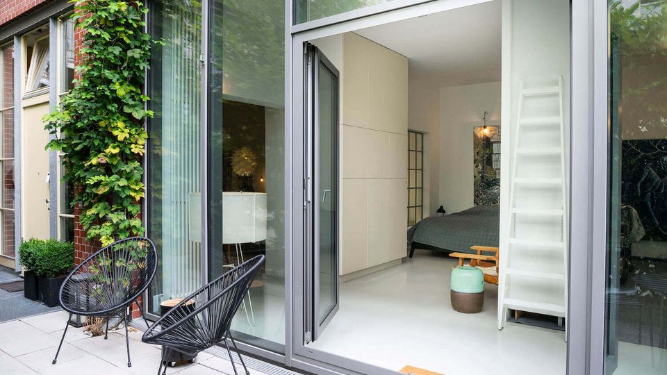 Ground-level patio of a modern brick-and-glass apartment with large sliding doors opening into a minimalist bedroom. The terrace has two black wire chairs and potted greenery, with climbing vines on the exterior wall and a white ladder-style shelving unit visible inside.