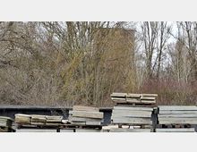 Gestapelte Betonplatten und Paletten auf einem Lagerplatz im Freien, davor eine dunkle Abdeckung oder Wand. Im Hintergrund stehen kahle Bäume und Sträucher, dahinter ragt ein großes, rechteckiges Gebäude teilweise verdeckt hervor.