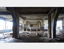 Innenraum einer verlassenen Fabrikhalle mit zerfallener Struktur. Sichtbar sind große Stützen, beschädigte Wände mit Graffiti, Trümmer und Schutt auf dem Boden sowie eingeschlagene Fenster, die Tageslicht einlassen.