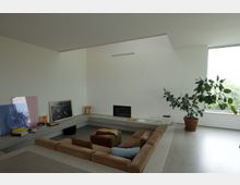 Moderner Wohnraum mit abgesenktem Sitzbereich, umgeben von einem beigen, gepolsterten Sofa. Die Wände sind weiß, mit minimaler Dekoration, darunter auf Regalen stehende gerahmte Bilder und Bücher. Eine große Zimmerpflanze steht neben einem Fenster, das viel natürliches Licht hereinlässt.