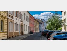 Straßenszene mit einer Reihe farbiger Altbauten, teils mit dekorativen Details, entlang einer schmalen Straße. Auf der rechten Seite parken Autos entlang eines Gehwegs; im Hintergrund sind Bäume und ein blauer Himmel mit Wolken zu sehen.