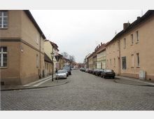 Straße mit Kopfsteinpflaster in einer Wohngegend, gesäumt von pastellfarbenen zweigeschossigen Häusern mit roten Ziegeldächern; parkende Autos stehen entlang beider Straßenseiten und ein einzelner Laternenpfahl ist auf dem Gehweg sichtbar.