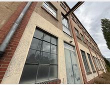 Schräg aufgenommene Fassade eines mehrstöckigen Industrie- oder Lagergebäudes mit rauem Putz, roten Ziegelpfeilern und großen, schwarz gerahmten Sprossenfenstern. Neben einem verwitterten Metalltor sind Regenfallrohre und eine kleine Außenlampe zu sehen, im Hintergrund Himmel und Bäume.