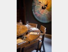 Runder Holztisch mit gedeckten Plätzen, darunter Gläser, Besteck und Menükarten, in einem modern eingerichteten Raum mit dunkler Wand; im Hintergrund ein großformatiges, abstraktes Wandbild mit bunten, radialen Mustern.