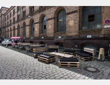 Backsteinfassade eines historischen Industriegebäudes mit großen, bogenförmigen Fenstern. Davor stehen Sitzmöbel aus Holzpaletten mit schwarzen Polstern, ein improvisierter Außenbereich mit Tischen und Stühlen sowie ein Verkaufsstand unter einem pinken Sonnendach auf einem gepflasterten Hof.