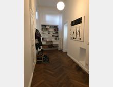 Flur mit dunklem Fischgrätparkettboden, weiß gestrichenen Wänden und einer minimalistisch gestalteten Deckenlampe. An der rechten Wand hängt ein abstraktes Gemälde, und am Ende des Flurs befindet sich ein Bücherregal mit diversen Büchern und Dekorationsgegenständen. Links steht eine Garderobe mit Jacken und Schuhen.