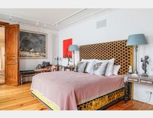 Schlafzimmer mit Holzboden, einem großen Bett mit einem dekorativen, wabenförmigen Kopfteil und Kissen mit Blumenmustern. Die Einrichtung umfasst einen rustikalen Holztürrahmen, eine kunstvolle Kommode, Lampen und Wanddekorationen, darunter ein großes Gemälde und eine rote abstrakte Leinwand.