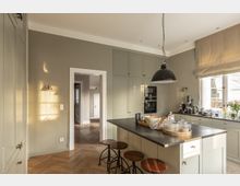 Helle, moderne Küche mit pastellfarbenen Schränken, einer schwarzen Kücheninsel mit Holzbarhockern und einem großen schwarzen Hängeleuchter. Rechts befindet sich ein Fenster mit beigefarbenem Rollvorhang, durch das Tageslicht einfällt, und im Hintergrund ist ein offener Durchgang zu einem weiteren Raum sichtbar.