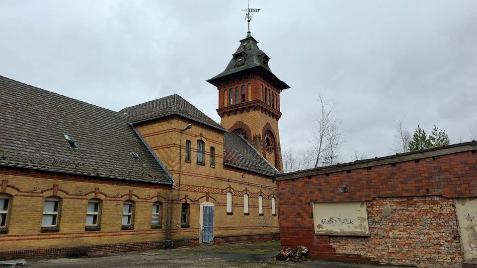 Backsteinbau mit langem, niedrigem Gebäudetrakt und einem hohen Turm mit dunkler Dachhaube und Wetterfahne. Im Vordergrund liegt ein asphaltierter Hof; rechts steht eine niedrige Mauer mit teils freigelegtem Mauerwerk unter grauem, bewölktem Himmel.