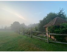 Ländliche Szene bei Nebel mit einer kleinen Hütte mit Ziegeldach, die von Pflanzen bedeckt ist, und einem Holzzaun, der einen Garten abgrenzt. Im Hintergrund sind Bäume und ein Bezug zur natürlichen Umgebung erkennbar, während der Himmel teils von Nebel bedeckt ist.