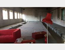 Ein großer, leerer Raum mit unverputzten Ziegelwänden, mehreren hohen Fenstern, die Tageslicht hereinlassen, und einem unvollständigen Betonboden mit eingelassenen Vertiefungen in der Mitte. Im Vordergrund befinden sich ein rotes Sofa, eine rote Stehlampe mit Fransen und ein roter Couchtisch mit einer Dekovase.