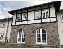 Fachwerkhaus mit weiß verputzten Wänden und dunklen Holzstreben im oberen Bereich. Unterer Bereich aus Natursteinmauerwerk mit zwei großen, weißen, bogenförmigen Fenstertüren; gepflasterter Außenbereich im Vordergrund.