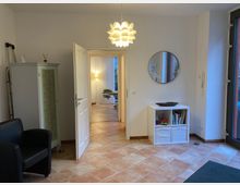 Heller Innenraum mit Fliesenboden, bestehend aus einem Warteraum mit einem schwarzen Sessel, einem weißen Schrank und einem Regal. Durch eine geöffnete Tür ist ein weiterer Raum mit einer Behandlungsliege sichtbar; eine moderne Lampe hängt von der Decke, und eine große Glastür zeigt nach draußen.