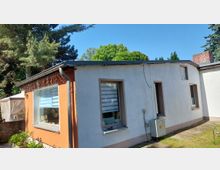 Anbau eines eingeschossigen Wohnhauses mit flachem Dach, orange-weißer Fassade und mehreren Fenstern mit Jalousien. Daneben verläuft ein schmaler Weg entlang der Wand, im Hintergrund stehen hohe Bäume und ein Gartenbereich.
