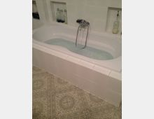 Weiße Badewanne mit eingelassenem Wasser, umgeben von weißen Wandfliesen und dekorativ gefliestem Boden in einem Bad. Im Hintergrund stehen Glasflaschen und Spenderflaschen in einer eingelassenen Nische.