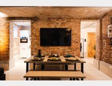 Wohnzimmer mit freigelegten Ziegelwänden und einem Gewölbedeckenmuster. Im Vordergrund steht ein gedeckter Esstisch mit Bänken, dahinter ein schwarzer Fernseher an der Wand; im Hintergrund sichtbar sind eine offene Küche und eine Holztür.