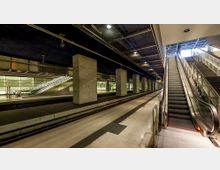 Unterirdischer Bahnhof mit beleuchteten Bahnsteigen, breiten Betonpfeilern und gläsernen Wänden im Hintergrund. Rechts führt eine Rolltreppe in eine obere Ebene, während links weitere Rolltreppen sichtbar sind.