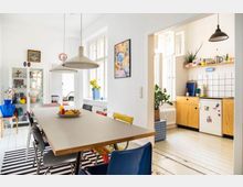 Esszimmer mit einem rechteckigen Holztisch und bunt gemischten Stühlen, dazu moderne Hängelampen. Im Hintergrund eine offene Küche mit weißen Fliesen, hölzernen Schränken und einer kleinen, dekorierten Arbeitsfläche.