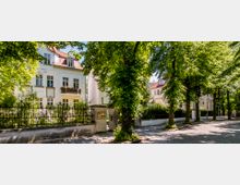 Straßenszene mit gepflastertem Gehweg, gesäumt von hohen, blattreichen Bäumen, die Schatten spenden. Links steht ein elegantes, helles Wohnhaus im klassischen Stil mit Balkonen und einem gepflegten Vorgarten, umgeben von einem schmiedeeisernen Zaun.