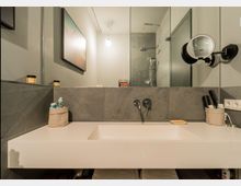 Modernes Badezimmer mit einem weißen Waschbecken auf einem schlichten Waschtisch, grauen Fliesen an der Wand und einer Glas-Duschkabine im Hintergrund. Auf dem Waschtisch stehen Pflegeprodukte und Pinsel in Behältern, und an der Wand ist ein runder, vergrößernder Spiegel angebracht.